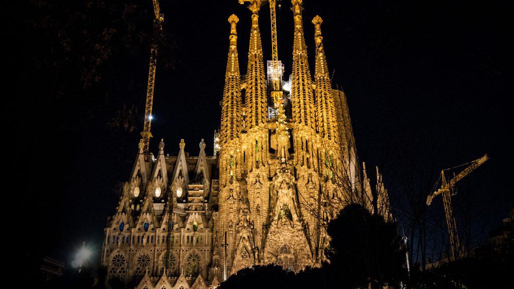 A night view of La Sagrada Familia in Barcelona, Spain, illuminated with construction cranes surrounding the iconic, intricate towers of the basilica. The golden lights highlight the architectural details against the dark sky.